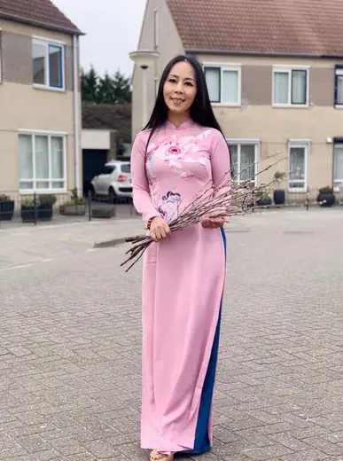 Một phụ nữ mặc áo dài hồng, quần xanh, cầm cành hoa đứng trên phố.