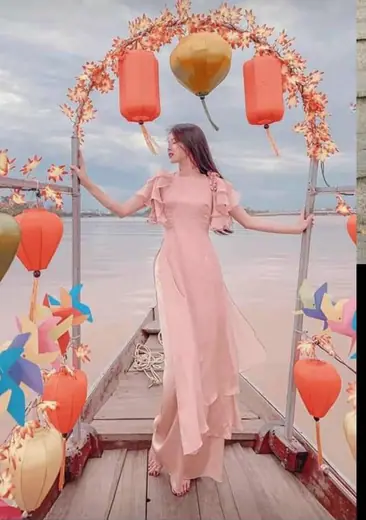 Một cô gái mặc áo dài hồng phấn đứng trên cầu gỗ treo đèn lồng bên sông.