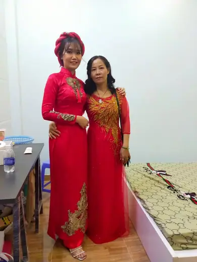 Hai phụ nữ mặc áo dài đỏ, một người đội mấn, đứng trong nhà.