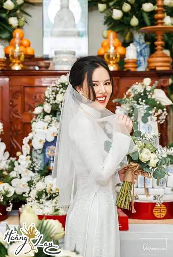 Cô dâu mặc áo dài ren trắng, đội khăn voan, ôm hoa trước bàn thờ gia tiên; ảnh có logo tiệm.