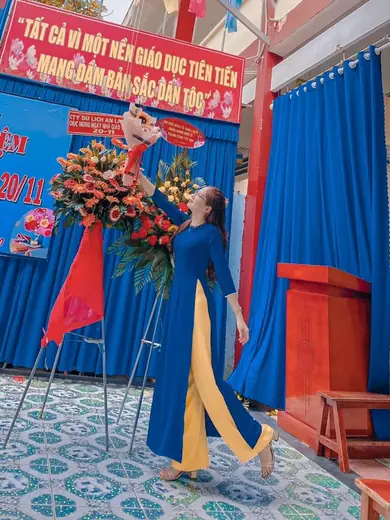 Một cô giáo mặc áo dài xanh dương, quần vàng, tạo dáng bên lẵng hoa ở sân trường.