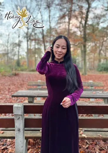 Một phụ nữ mặc áo dài nhung tím đứng bên dãy bàn ghế gỗ giữa rừng thu.