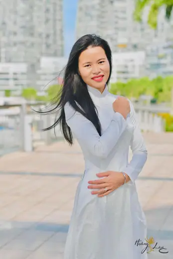 Một phụ nữ mặc áo dài trắng đứng trên lối đi ven sông, phía sau là các toà nhà cao tầng.