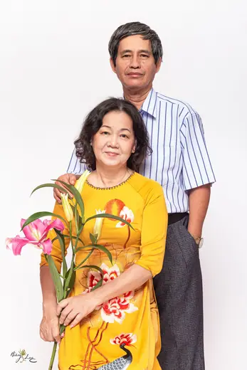 Một cặp vợ chồng lớn tuổi chụp trong phòng chụp, người vợ mặc áo dài vàng vẽ hoa sen và cầm hoa.