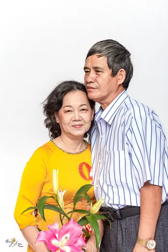 Người vợ mặc áo dài vàng tựa vào chồng, tay cầm hoa ly.
