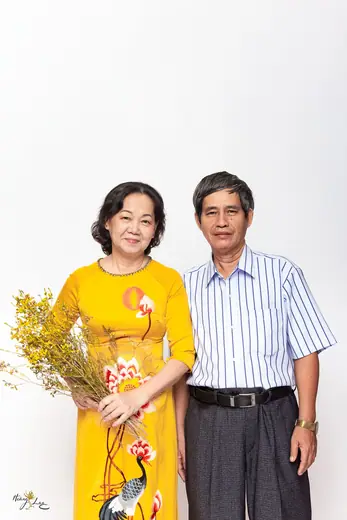 Cặp vợ chồng lớn tuổi đứng cạnh nhau, người vợ mặc áo dài vàng vẽ chim hạc.