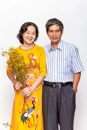 Người vợ mặc áo dài vàng vẽ chim hạc ôm bó hoa, đứng bên chồng.