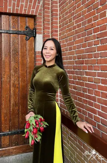 Một phụ nữ mặc áo dài nhung xanh rêu, quần vàng, đứng bên cánh cửa gỗ.
