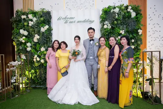 Cô dâu mặc váy cưới và chú rể mặc vest chụp cùng người thân mặc áo dài trước phông tiệc cưới.