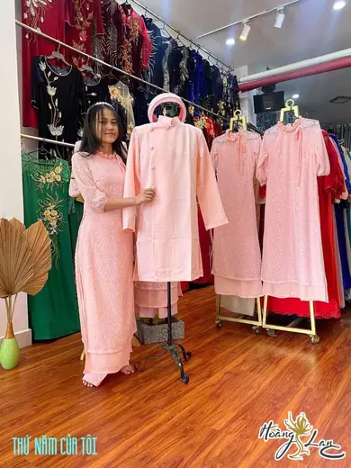 Một cô gái mặc áo dài hồng đứng bên chiếc áo dài nam màu hồng trên ma-nơ-canh trong tiệm, ảnh có logo tiệm.