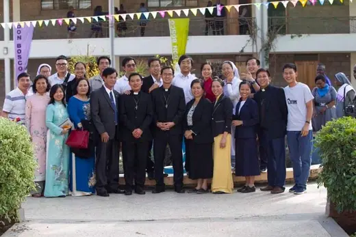 Một đoàn đông người, có cả tu sĩ, chụp chung trước toà nhà treo dây cờ; vài phụ nữ mặc áo dài.