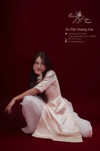 Một cô gái mặc áo dài ren trắng ngồi trên nền đỏ sẫm, ảnh có logo và địa chỉ tiệm.