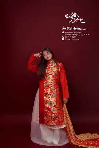 Một cô gái mặc áo dài gấm đỏ vàng khoác ngoài lớp áo trắng, choàng khăn đỏ, trên nền đỏ sẫm; ảnh có logo tiệm.