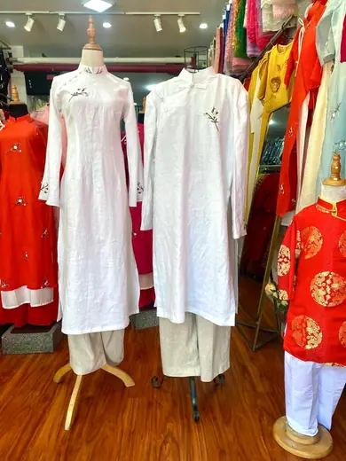 Hai chiếc áo dài vải đũi trắng thêu hoa, một nữ một nam, trên ma-nơ-canh trong tiệm.