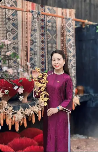 Một phụ nữ mặc áo dài tím hồng đứng bên bó chổi đót đỏ và hoa khô.