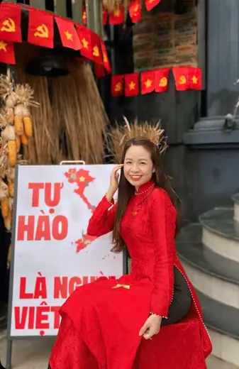 Một phụ nữ mặc áo dài đỏ ngồi trước góc trang trí cờ đỏ sao vàng.