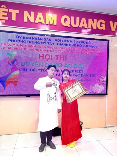 Một người đàn ông mặc áo dài trắng và một phụ nữ mặc áo dài đỏ cầm giấy khen trước màn hình hội thi Duyên dáng Áo dài.